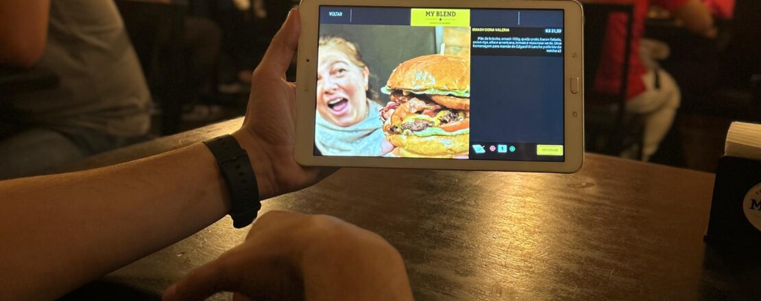 Cliente utilizando um tablet com o cardápio digital para fazer um pedido em uma hamburgueria, destacando a imagem de um hambúrguer suculento e as opções disponíveis no menu.